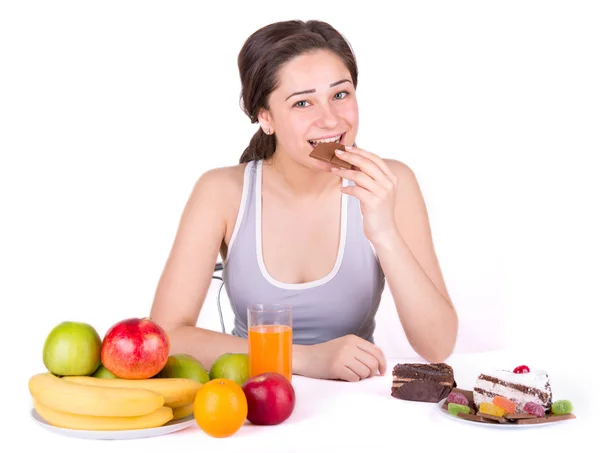 Девушка сидит рядом фрукты и сладости и кусает шоколад — стоковое фото