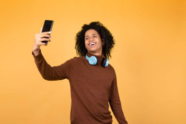 Neşeli afro genç adam telefonda selfie çekiyor. Sarı arka planda gelişigüzel giyinmiş Afrikalı bir gencin fotoğrafı..