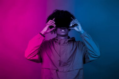 VR gözlüklü afro çocuk. Sanal gerçekliğe dalmış genç bir afro elleriyle hareket ediyor. Mavi ve kırmızı ışık bir çocuğun üzerine düşüyor..