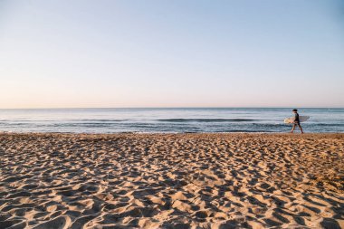 Genç Afro Latin adam deniz kıyısında sörf tahtasıyla yürüyor.