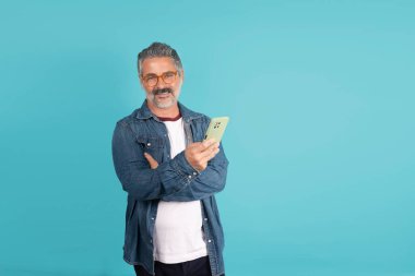 Olgun bir adam kollarını kavuşturarak akıllı telefonu tutarken gülümsüyor.