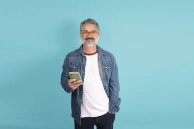 Gülümseyen, elinde cep telefonu olan, mavi arka planda giyinmiş olgun bir adam.