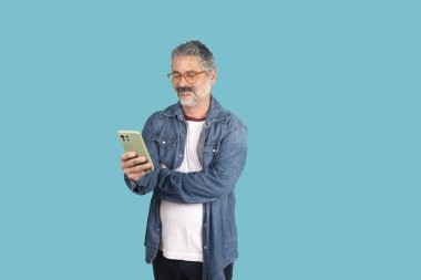Turkuaz arka planda cep telefonuyla etkileşime girerken gülümseyen olgun bir adam