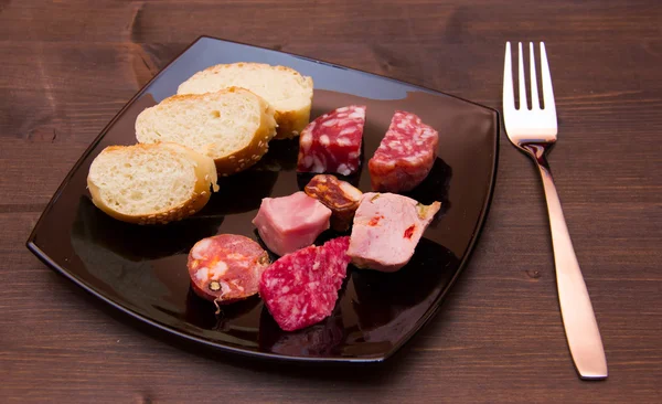 Trozos de salami y rebanadas de pan en plato sobre madera — Foto de Stock