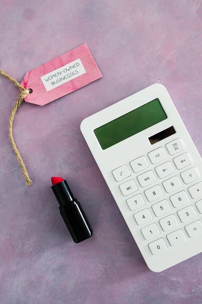 women-owned business tag with calculator and lipstick on pink desk, supporting equality and equal opportunities