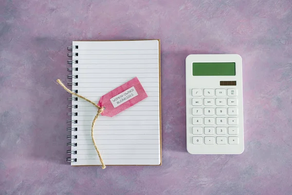 women-owned business tag with calculator and notepad on pink desk, supporting equality and equal opportunities