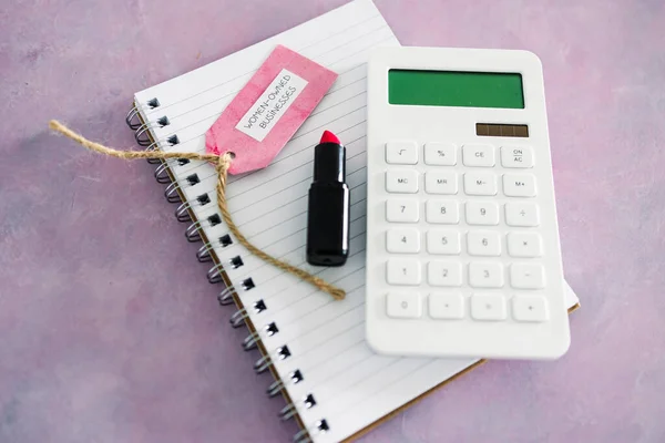 women-owned business tag with calculator notepad and lipstick on pink desk, supporting equality and equal opportunities