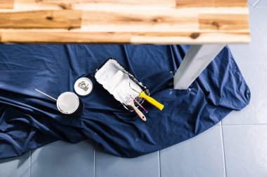 Boya tepsisi silindirli bir tencere beyaz boya ve mavi çarşafın üstünde fırçalar. Tabloyu boyamaya hazır. Boya kutusuyla sığ bir alanda çekilmiş.