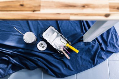 Boya tepsisi silindirli bir tencere beyaz boya ve mavi çarşafın üstünde fırçalar. Tabloyu boyamaya hazır. Boya kutusuyla sığ bir alanda çekilmiş.