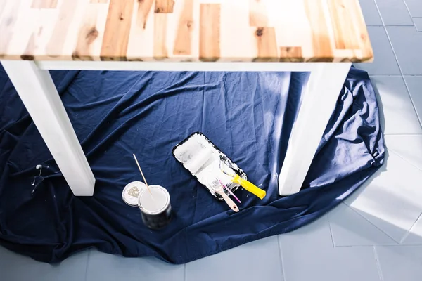 pot of white paint with paint tray roller and brushes on top of blue drop sheet ready to paint a table, shot at shallow depth of field with paint can text blurred