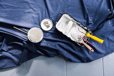 Boya tepsisi silindirli bir tencere beyaz boya ve mavi çarşafın üzerindeki fırçalar boyaya hazır, boya kutusuyla bulanık bir şekilde sığ alan derinliğinde çekiliyor.