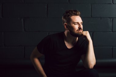 Handsome bearded men in black shirt thinking about his life near brick wall