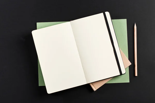 Open blank notepad with empty white pages laying above book with pencil on black background. ideal for text — ストック写真