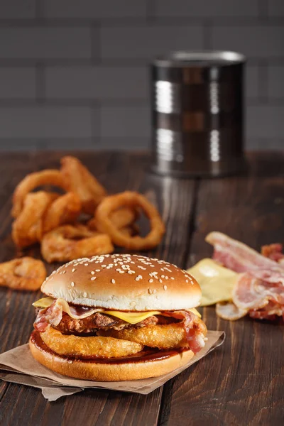 Smaczny grill burger z boczkiem i cebulą pierścienie na drewnianym stole w kuchni. szablon szybkiej obsługi. niezdrowe jedzenie — Zdjęcie stockowe