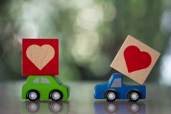 Couple of wooden toy cars delivering wooden hearts for Valentine\'s day on nature blur background. Concept of delivering love