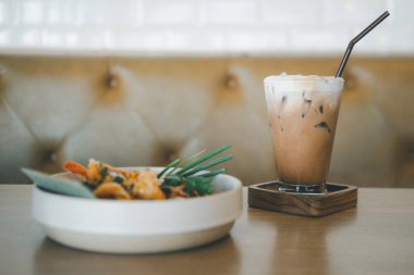 Kafedeki ahşap masada Tayland yemeğiyle servis edilen uzun bardakta buzlu kahve.