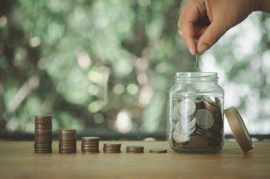 Hand holding coin put into money saving bottle on wood table against nature background.  Money saving idea to invest in the future.