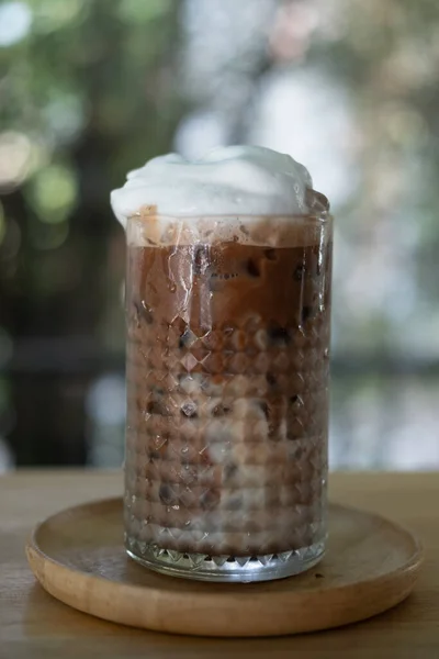 A glass of iced coffee mocha with foam milk on top on wood table