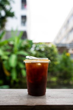 Closeup of take away plastic cup of iced black coffee Americano on wooden table with green nature background.
