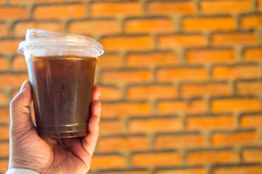 El ele tutuşmuş buzlu kahve ya da plastik bardakta Amerikan kahvesi, tuğla arka planda.
