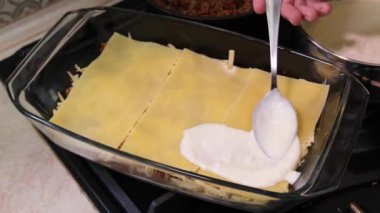 A woman in the kitchen prepares lasagna: pasta of lasagna, adds cheese, red minced meat sauce (bolognese) and white sauce (bechamel). Cooking lasagna