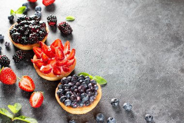 Gri arka planda kremalı meyveli tartlar. Yaz meyveli kek. Çilekli tart, yaban mersini, böğürtlen. Seçici odaklanma. Üst görünüm.