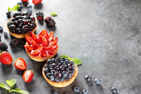 Gri arka planda kremalı meyveli tartlar. Yaz meyveli kek. Çilekli tart, yaban mersini, böğürtlen. Seçici odaklanma. Üst görünüm.