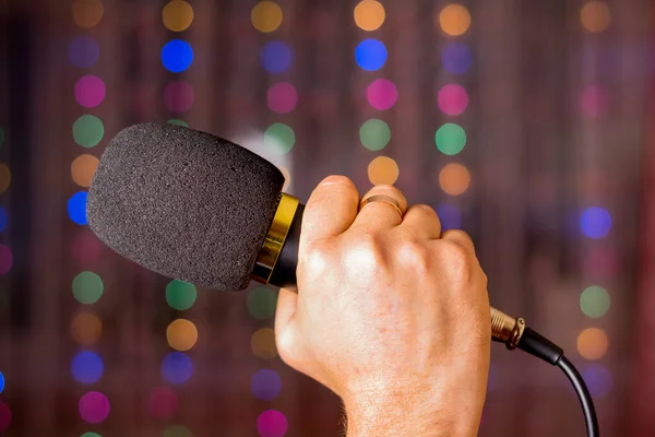 Microphone in hand. Interview. Karaoke. A man's hand holds a microphone against the background of blurry garlands