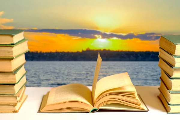 A stack of books and an open book against the backdrop of a sunset over the river. Reading visualization concept