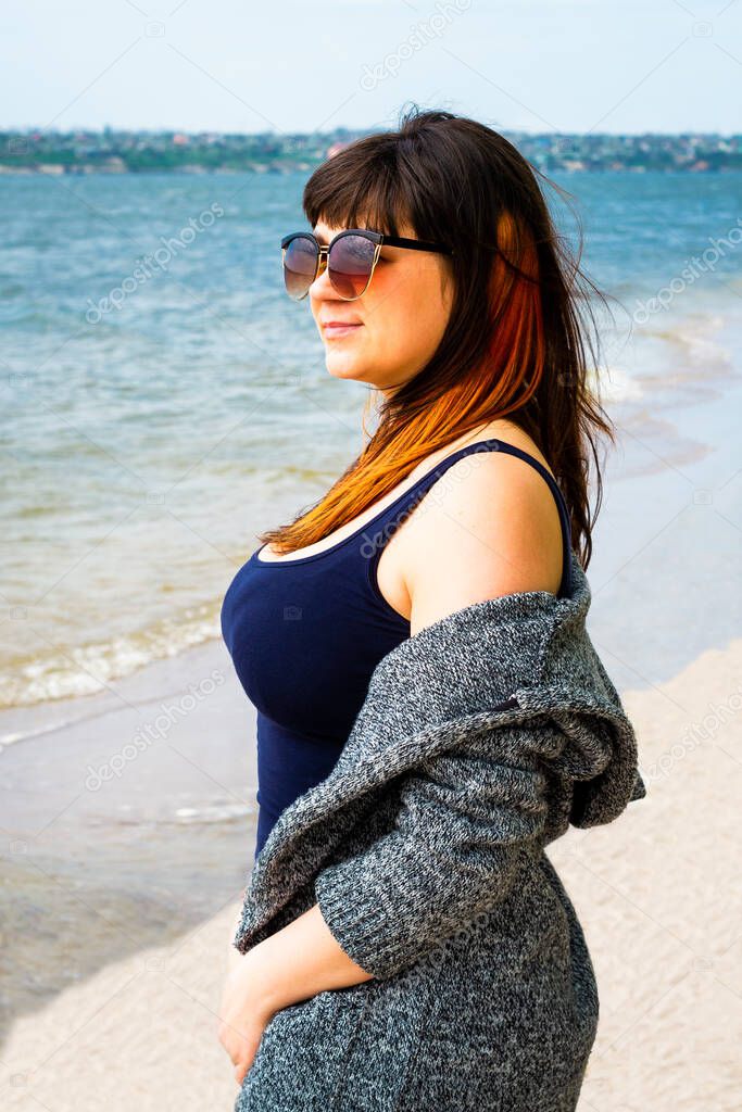 A young woman in sunglasses with auburn hair, in a blue T-shirt and a gray cardigan looks into the distance against the background of the river