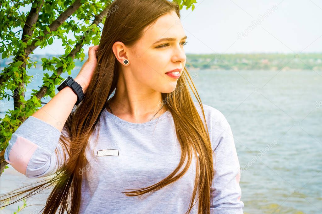 A young brunette in a gray sweatshirt straightens her hair and looks to the side against a background of a green tree and a river