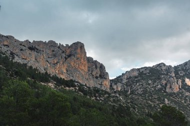 İspanya 'nın Puig Campana bölgesinde, kasvetli bir gökyüzü altında yoğun çam ormanlarının üzerinde görkemli kayalık kayalıklar yükselir. Doğa, seyahat ve macera temaları için mükemmel.