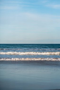Açık mavi gökyüzü altında, boş kumlu bir sahilde, arka plan, rahatlama temaları veya kıyı tasarımı projeleri için ideal, yumuşak okyanus dalgaları olan en az deniz manzarası..
