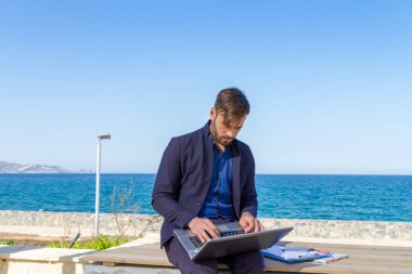 Okyanus kenarında cep telefonu ve dizüstü bilgisayar kullanan genç bir adam.