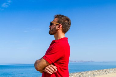 Dinamik, hırslı, başarılı, kırmızı tişörtlü, sakallı genç, mavi gökyüzü arka planında güneş gözlüklü. Özgüven yaşam tarzı kavramı