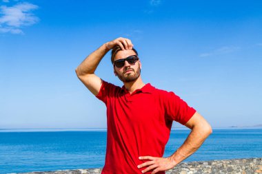 Dinamik, hırslı, başarılı, kırmızı tişörtlü, sakallı genç, mavi gökyüzü arka planında güneş gözlüklü. Özgüven yaşam tarzı kavramı