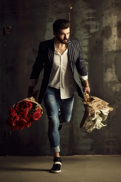 Handsome young man in smart jacket with bouquet of flowers  poses in front of the  wall — Stock Photo, Image