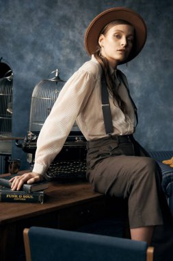 Eski tarz bir kızın portre fotoğrafı. İç stüdyoda poz veriyor. Pantolon, gömlek, pantolon askısı ve şapka giyiyor.