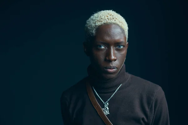 portrait of a dark-skinned handsome guy with white hair and blue eyes, who looks into the camera with a serious expression, he is dressed in a brown sweater over which hangs a silver chain