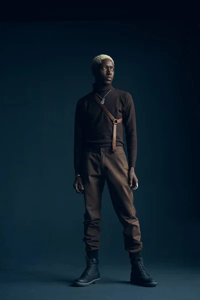 full-length photo in the studio of a dark-skinned handsome guy with white hair and blue eyes standing on a dark background with a serious facial expression in brown clothes and silver accessories