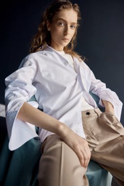 Stüdyoda, koyu renk arka planda, büyük mavi gözlü, uzun dalgalı saçlı güzel bir kızın portresi. Altında bej pantolon ve beyaz bir pamuk gömlek giyiyor.