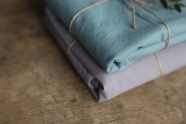 2 doğal ve ekolojik kumaşın gri ve turkuaz renklerle yıkanmış fotoğrafı üzerinde çuval ipliği ve yeşil bitkiyle geri sarılmış.