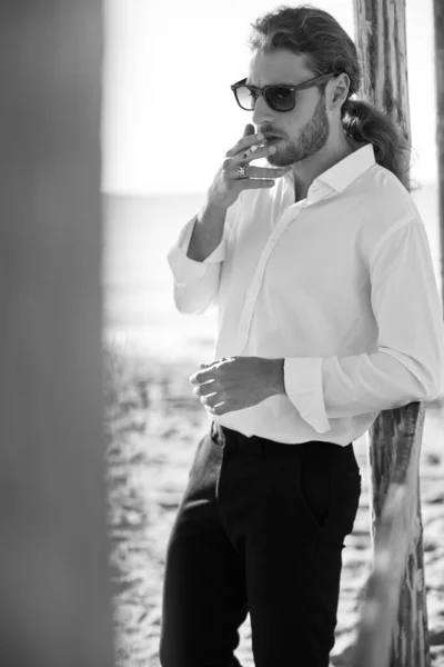 黒と白の写真は成長の真っただ中に立って海のそばにタバコを灯しているスタイリッシュな男 — ストック写真
