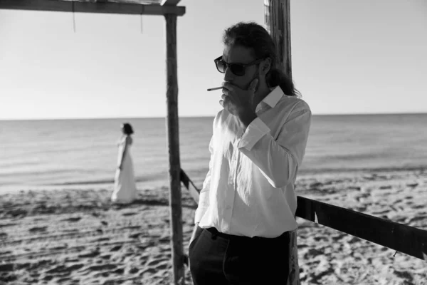 black and white photo a guy with long hair in a white shirt standing on the sand by the sea and smoking a cigarette and a girl in a white dress on the background