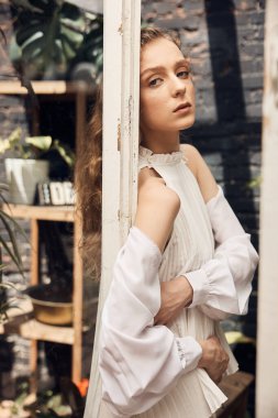 Mavi gözlü, uzun kıvırcık saçlı, beyaz elbiseli, siyah tuğlalı bir duvarın arkasında duran genç bir kız.