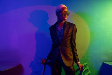 Siyah takım elbiseli esmer adam. Metal süslemelerle. O, siyah gözlük takıyor. Mor ve yeşil ışık.