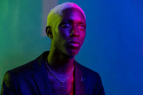 dark-skinned guy with a white face looking at a purple light. Dressed in a black jacket and silver jewelry around his neck