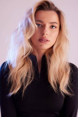 Sarışın, kıvırcık saçlı, kameraya bakıyor. Siyah bir kazak giymişti.