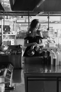 Siyah elbiseli, uzun saçlı, harika bir kadının siyah beyaz fotoğrafı. Yan tarafa bakıp gülüyor ve elinde bir tava tutuyor.