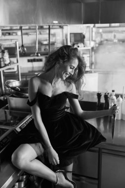 Masanın etrafında kısa, şık bir siyah elbiseyle duran bir kızın fotoğrafı. Elinde metal bir bıçak tutuyor ve gülümsüyor.